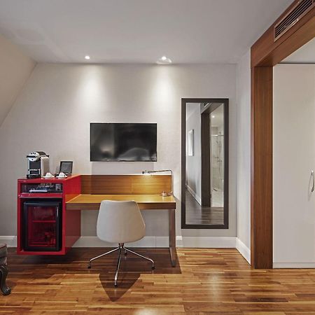 ไททานิค เกนดาร์เมนมาร์ค เบอร์ลิน โฮเทล Hotel ภายนอก รูปภาพ The photo depicts a modern, well-lit room featuring a small workspace. On the right side, there is a compact desk with a red and wooden design, accompanied by a comfortable white chair with wheels. Above the desk is a flat-screen TV mounted on the wa