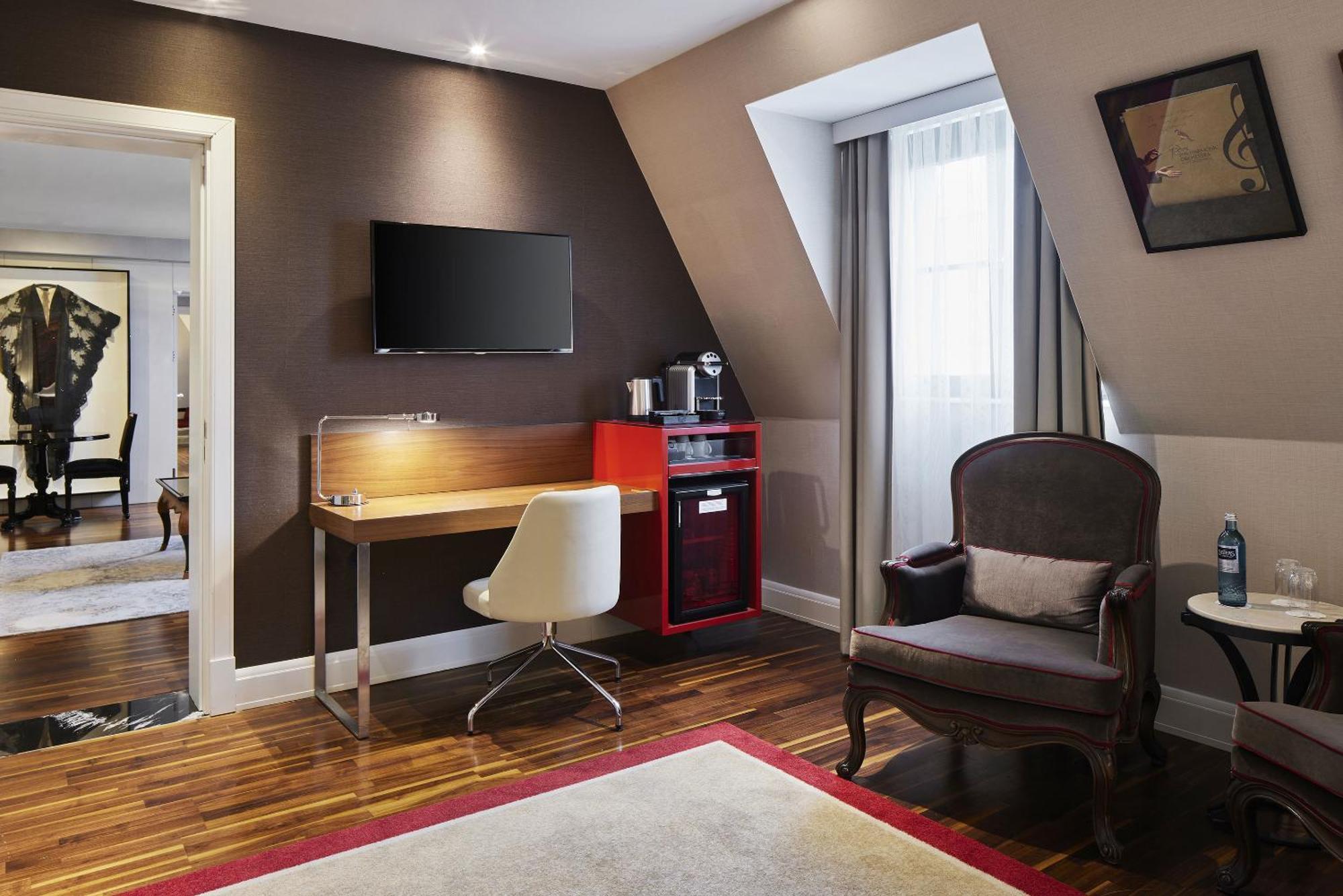 ไททานิค เกนดาร์เมนมาร์ค เบอร์ลิน โฮเทล Hotel ภายนอก รูปภาพ The photo depicts a modern hotel room or a cozy living area. It features a sloped ceiling with soft lighting. On one wall, there is a flat-screen TV mounted above a wooden desk with a sleek, white chair. To the side, there is a small red minibar or c
