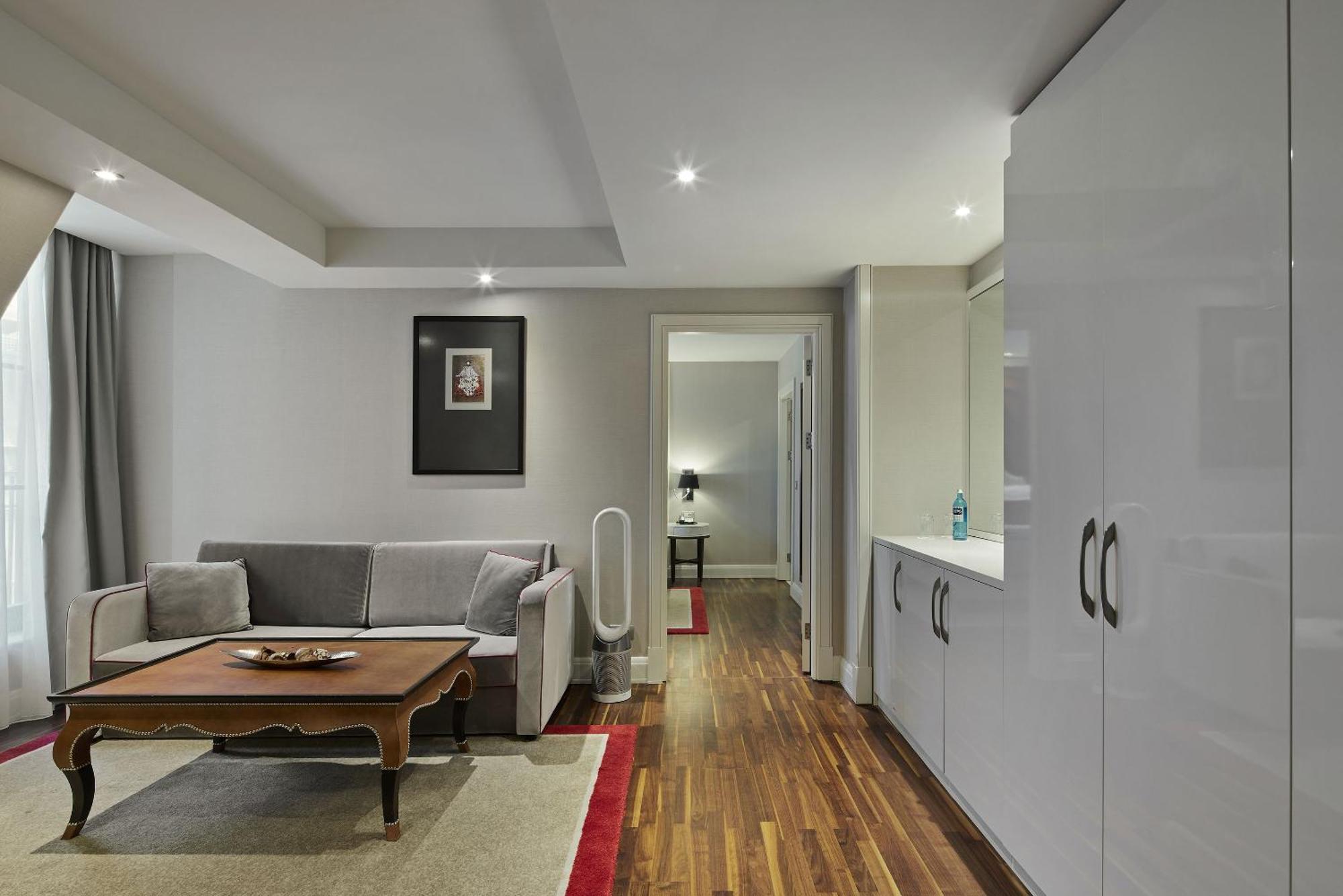 ไททานิค เกนดาร์เมนมาร์ค เบอร์ลิน โฮเทล Hotel ภายนอก รูปภาพ The photo shows a modern living room area featuring a gray sofa and a wooden coffee table. The floor is made of polished wood, and there is a light-colored rug underneath the table. A framed picture hangs on the wall to the left, and there are curtai