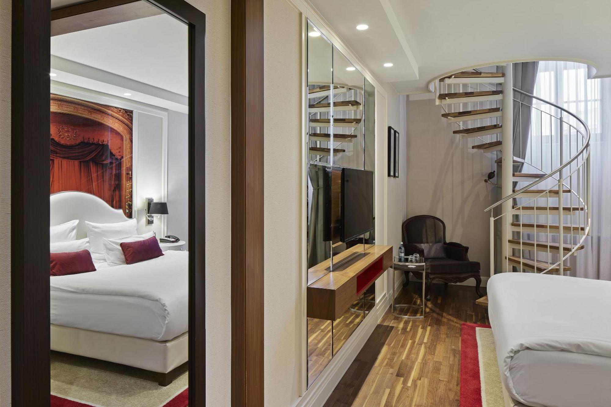 ไททานิค เกนดาร์เมนมาร์ค เบอร์ลิน โฮเทล Hotel ภายนอก รูปภาพ The photo shows an interior space, likely a hotel room or a suite. On one side, there is a bedroom with a large bed, decorative pillows, and artwork above the bed. The opposite side features a seating area with a dark chair and a small table, along w