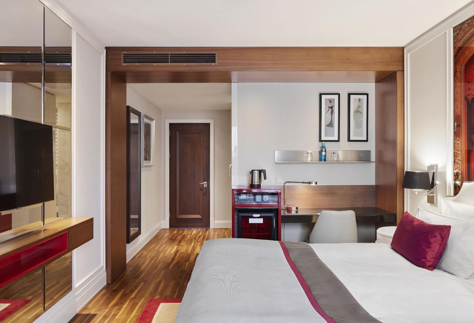 ไททานิค เกนดาร์เมนมาร์ค เบอร์ลิน โฮเทล Hotel ภายนอก รูปภาพ The photo shows a modern hotel room featuring a king-sized bed with gray bedding and decorative pillows. There's a sleek wooden wall with a mounted television opposite the bed. To the side, there is a work desk with a chair and a small refrigerator o